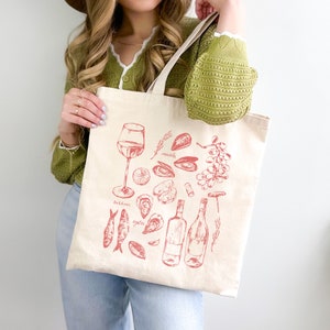 Op de afbeelding: Een witte canvas tote bag met een rode illustratie van wijn, zeevruchten en andere etenswaren. De tekst op de tas is "Sardines", "oesters" en "mosselen".