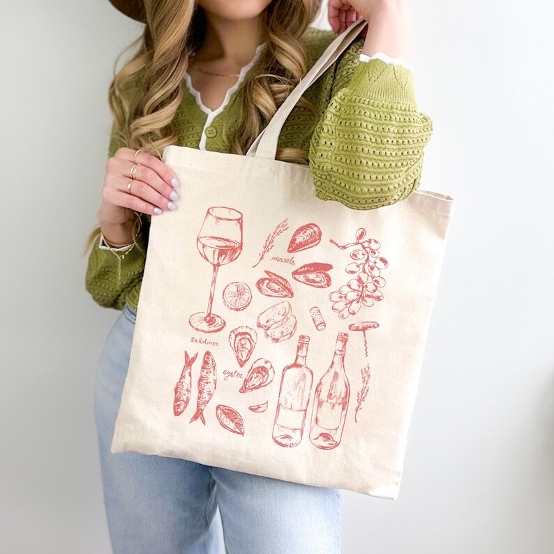 Op de afbeelding: Een witte canvas tote bag met een rode illustratie van wijn, zeevruchten en andere etenswaren. De tekst op de tas is "Sardines", "oesters" en "mosselen".