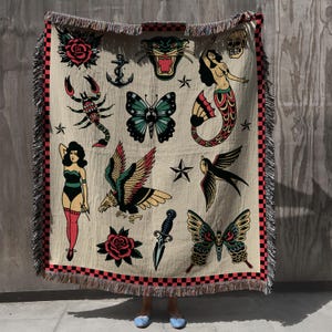 May include: A woven tapestry throw blanket with a cream background and red, black, green, and blue traditional tattoo-style imagery. The imagery includes a scorpion, an eagle, a butterfly, a dagger, a mermaid, a swallow, a rose, an anchor, and a skull.