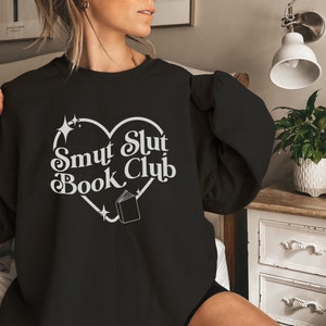 以下が含まれることがあります： 白いハート型のデザインに「Smut Slut Book Club」の文字が入った黒いスウェットシャツ。デザインには本のイラストと星のアクセントが含まれています。カジュアルな衣服。