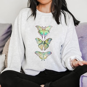 May include: A light gray sweatshirt with a design of three iridescent butterflies in a vertical row. The butterflies are green, blue, and yellow.