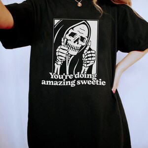 May include: Black t-shirt featuring a graphic of a skeleton wearing a hooded robe and giving a thumbs up. The text "You're doing amazing sweetie" is printed below the graphic.