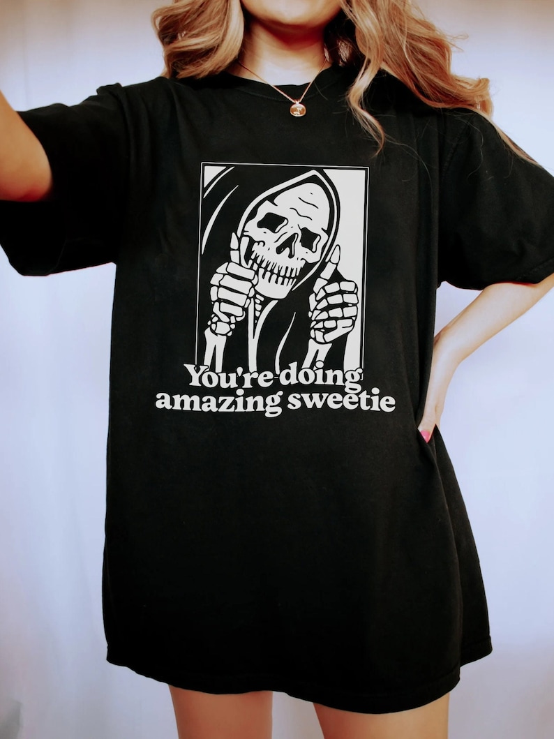 May include: Black t-shirt featuring a graphic of a skeleton wearing a hooded robe and giving a thumbs up. The text "You're doing amazing sweetie" is printed below the graphic.