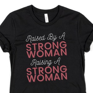 Puede incluir: Camiseta negra con el texto "Raised By A STRONG WOMAN Raising A STRONG WOMAN" en letras blancas y rosas.