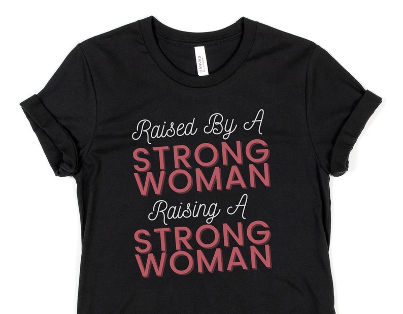 Puede incluir: Camiseta negra con el texto "Raised By A STRONG WOMAN Raising A STRONG WOMAN" en letras blancas y rosas.