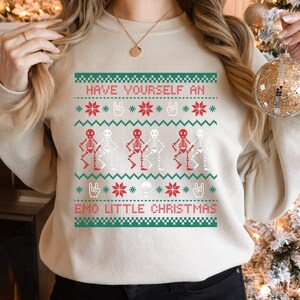 K&ouml;nnte beinhalten: Ein cremefarbener Pullover mit einem festlichen Design mit Skeletten in Rot und Wei&szlig;, Schneeflocken und dem Text "Have Yourself an Emo Little Christmas".