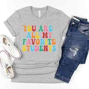 May include: A grey t-shirt with the text "You are all my favourite students" in colourful letters. The shirt is paired with blue jeans and white trainers.
