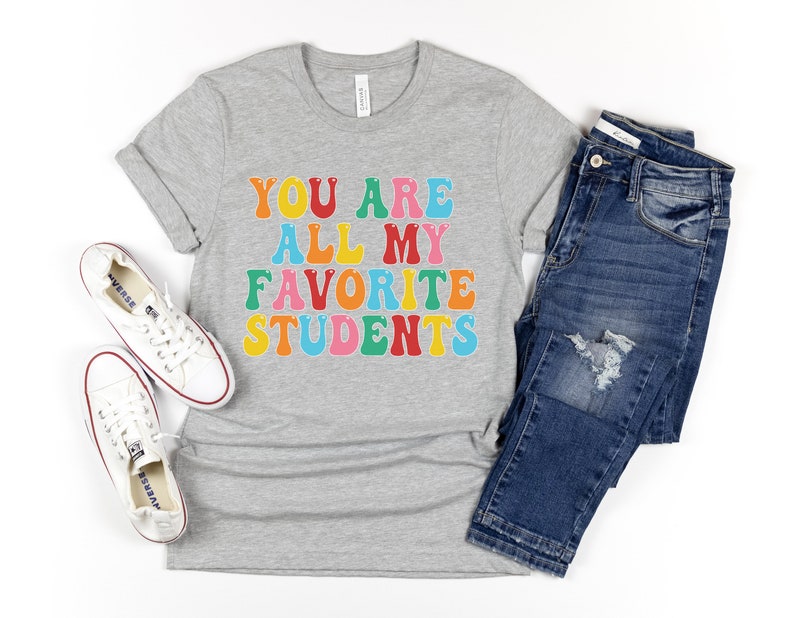 May include: A grey t-shirt with the text "You are all my favourite students" in colourful letters. The shirt is paired with blue jeans and white trainers.