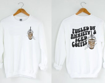 May include: White long-sleeve sweatshirt with the text "Fueled by Anxiety & Iced Coffee" in black and a graphic of a skeleton hand holding a cup of iced coffee on the back. The front of the sweatshirt has a small graphic of a cup of iced coffee.
