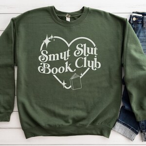 以下が含まれることがあります： オリーブグリーンのスウェットシャツ。白いハート型のデザインで「Smut Slut Book Club」の文字がプリントされています。デザインには本と星が含まれています。青いジーンズが見えます。