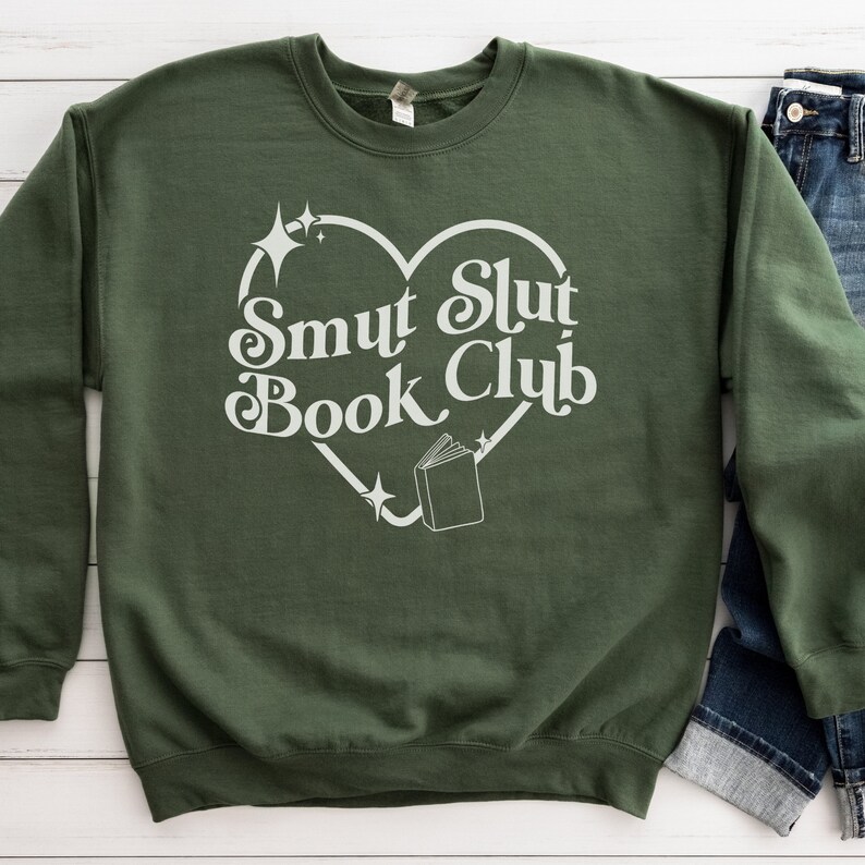 以下が含まれることがあります： オリーブグリーンのスウェットシャツ。白いハート型のデザインで「Smut Slut Book Club」の文字がプリントされています。デザインには本と星が含まれています。青いジーンズが見えます。