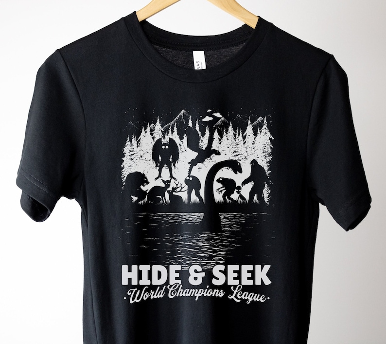 May include: Black t-shirt with a white graphic of various mythical creatures playing hide and seek in a forest. The text "Hide & Seek. World Champions League." is printed below the graphic.