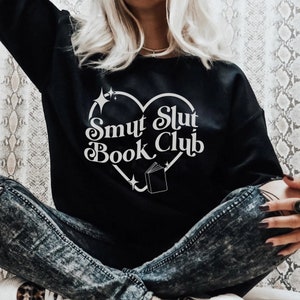 以下が含まれることがあります： 白いハート型のデザインに「Smut Slut Book Club」の文字が入った黒いスウェットシャツ。デザインには本と星のアクセントが含まれています。スウェットシャツはカジュアルなスタイルです。