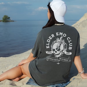 May include: A person wearing a dark gray t-shirt with a white skull graphic and the text "Elder Emo Club" on the back. The shirt has the text "Stay Sad" on a banner above the skull and "Happiness Fades Emo Is Forever" on a banner below the skull. The person is wearing a white baseball cap and sitting on a sandy beach.
