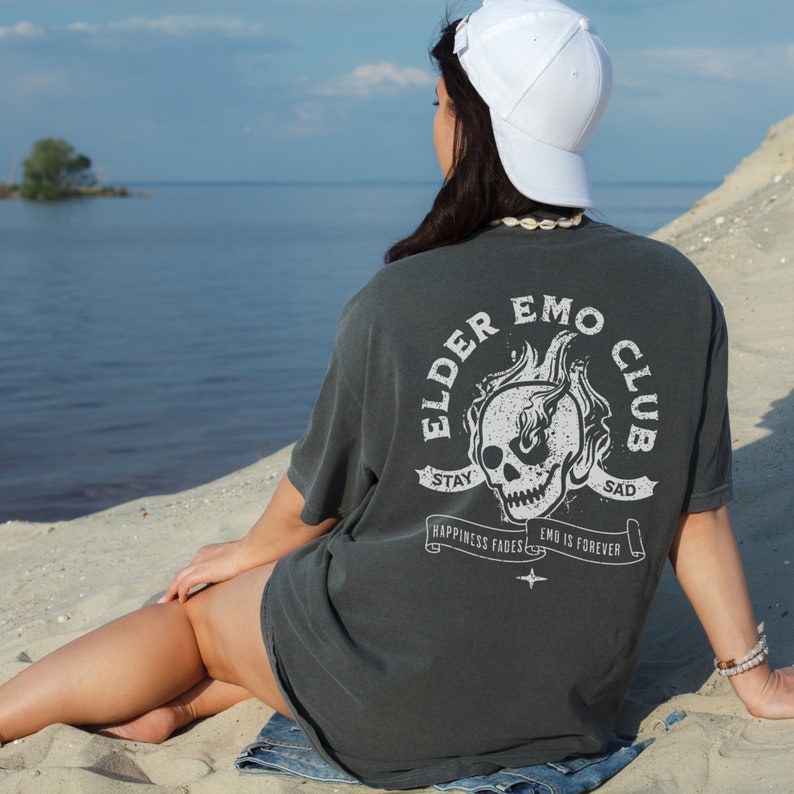 May include: A person wearing a dark gray t-shirt with a white skull graphic and the text "Elder Emo Club" on the back. The shirt has the text "Stay Sad" on a banner above the skull and "Happiness Fades Emo Is Forever" on a banner below the skull. The person is wearing a white baseball cap and sitting on a sandy beach.