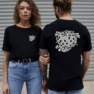 May include: Two people wearing black t-shirts. The person on the right has a white graphic on the back of their shirt that says "All Panic, No Disco" with a disco ball and skeleton hands.