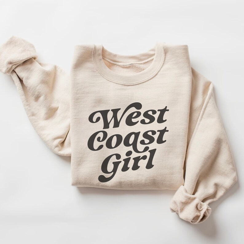 May include: A cream-colored sweatshirt with the words "West Coast Girl" printed in a dark gray, retro font. The sleeves are rolled up, and the sweatshirt is folded neatly on a white surface. The overall style is casual and comfortable.