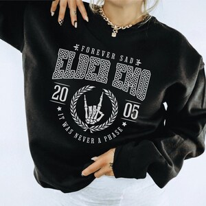 以下が含まれることがあります： 「Forever Sad Elder Emo 20 05 It Was Never A Phase」と白で書かれたグラフィックプリントが施された黒いスウェットシャツ。グラフィックには、市松模様、花輪、ロックンロールのハンドジェスチャーをするスケルトンの手が入っています。