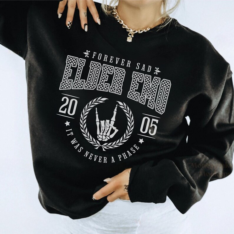 以下が含まれることがあります： 「Forever Sad Elder Emo 20 05 It Was Never A Phase」と白で書かれたグラフィックプリントが施された黒いスウェットシャツ。グラフィックには、市松模様、花輪、ロックンロールのハンドジェスチャーをするスケルトンの手が入っています。
