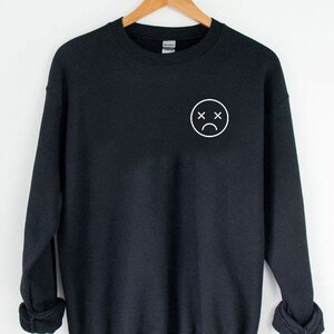 May include: Black sweatshirt with a white outline of a sad face with an X for eyes.