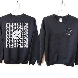 May include: Two black sweatshirts with white graphic designs. One sweatshirt has the text "STILL EMO" repeated in a wavy font. The other sweatshirt has a simple white sad face with an X for eyes.