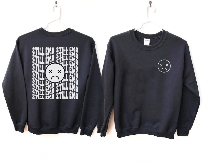 May include: Two black sweatshirts with white graphic designs. One sweatshirt has the text "STILL EMO" repeated in a wavy font. The other sweatshirt has a simple white sad face with an X for eyes.