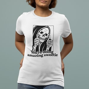 May include: White t-shirt featuring a black and white illustration of the Grim Reaper giving a thumbs up. The text "You're doing amazing sweetie" is printed below the illustration.