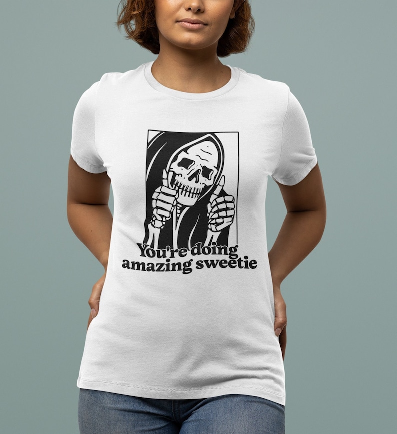 May include: White t-shirt featuring a black and white illustration of the Grim Reaper giving a thumbs up. The text "You're doing amazing sweetie" is printed below the illustration.