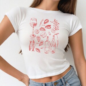 May include: White t-shirt with a red graphic design of various seafood, including oysters, mussels, sardines, and wine bottles. The text "oysters", "mussels", "sardines" is included in the design.