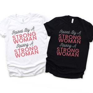 Puede incluir: Dos camisetas, una blanca y otra negra, con el texto "Raised By A Strong Woman Raising A Strong Woman" en letras negras y rosas.