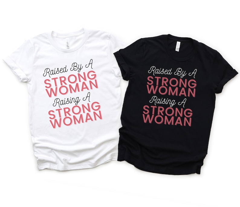 Puede incluir: Dos camisetas, una blanca y otra negra, con el texto "Raised By A Strong Woman Raising A Strong Woman" en letras negras y rosas.