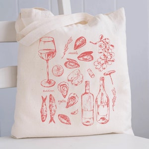 Op de afbeelding: Een witte canvas draagtas met een rood ontwerp van verschillende zeevruchten, wijn en wijnaccessoires. Het ontwerp omvat mosselen, oesters, sardines, een glas wijn, twee flessen wijn, een kurkentrekker en druiven. De tekst op de tas luidt "mosselen", "sardientjes" en "oester".
