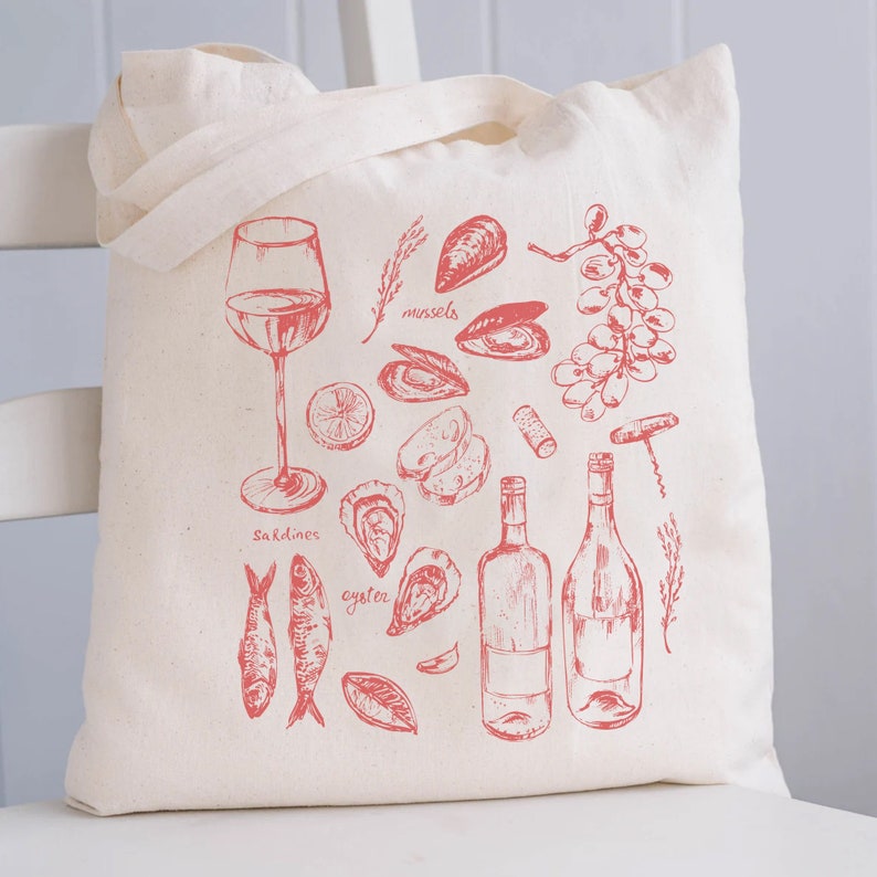Op de afbeelding: Een witte canvas draagtas met een rood ontwerp van verschillende zeevruchten, wijn en wijnaccessoires. Het ontwerp omvat mosselen, oesters, sardines, een glas wijn, twee flessen wijn, een kurkentrekker en druiven. De tekst op de tas luidt "mosselen", "sardientjes" en "oester".