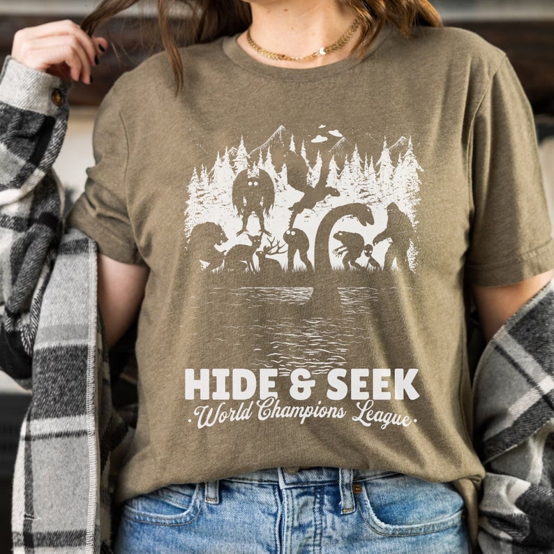 May include: A green t-shirt with a white graphic of various mythical creatures, including a monster, a sasquatch, and a loch ness monster. The text "Hide & Seek World Champions League" is printed below the graphic.