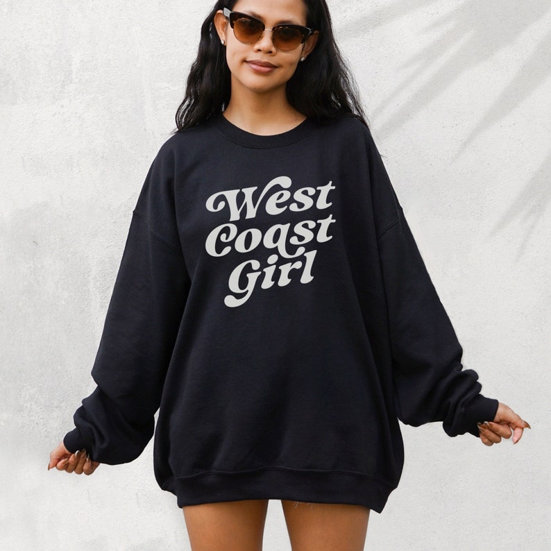 May include: Black sweatshirt with the text "West Coast Girl" in a white, retro font. The sweatshirt has long sleeves and a relaxed fit. The image shows a person wearing sunglasses.