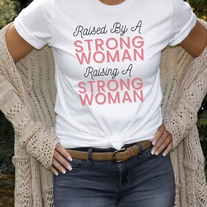 Puede incluir: Una camiseta blanca con el texto "Raised By A Strong Woman Raising A Strong Woman" en letras negras y rosas.