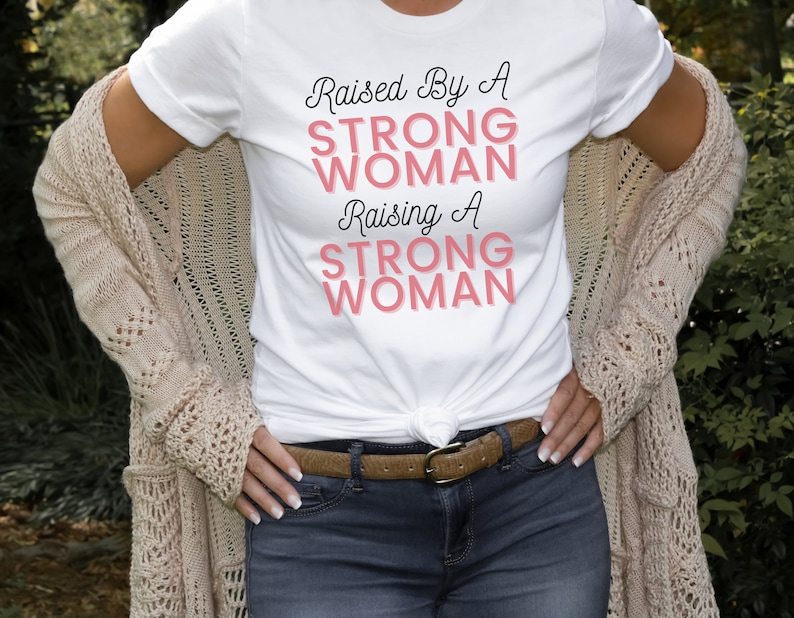 Puede incluir: Una camiseta blanca con el texto "Raised By A Strong Woman Raising A Strong Woman" en letras negras y rosas.