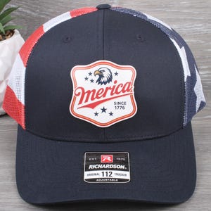 May include: A navy blue trucker hat featuring a red, white, and blue flag pattern on the mesh back panels. The front panel displays a shield-shaped patch with an eagle graphic and the text Merica SINCE 1776. A black Richardson 112 sticker is attached.