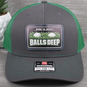 May include: A gray and green trucker hat with a patch that reads "BORN TO BOGEY! BALLS DEEP" featuring two golf balls in grass. The hat is adjustable and has a Richardson 112 Trucker tag.