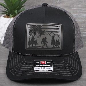 May include: Black and gray trucker hat with a Bigfoot and American flag patch. The patch features a mountain range, trees, and a silhouette of Bigfoot. The hat has a curved brim and an adjustable strap.