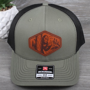 May include: A green and black trucker hat with a patch featuring a Bigfoot illustration in an orange and brown design. The hat has a curved brim and an adjustable strap. The patch reads "RICHARDSON 112 TRUCKER".