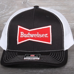 May include: A black and white trucker hat with a red Budweiser logo. The hat is made by Richardson and has an adjustable strap.