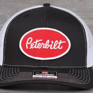 May include: Black and white trucker hat with a red oval patch that says "Peterbilt" in white cursive lettering. The hat is adjustable and has a white mesh back.