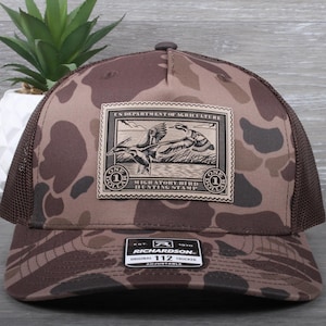 May include: A brown and tan camouflage trucker hat with a brown leather patch that says "U.S. Department of Agriculture Migratory Bird Hunting Stamp". The patch features a black and white illustration of two ducks in flight. The hat is adjustable and has a black mesh back.