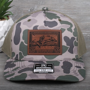 May include: A camouflage trucker hat with a brown leather patch featuring a vintage duck hunting stamp. The patch reads "U.S. Department of Agriculture Migratory Bird Hunting Stamp". The hat is adjustable and has a mesh back.