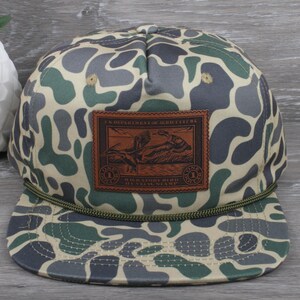 May include: A camouflage patterned baseball cap with a brown leather patch featuring a vintage duck hunting stamp. The stamp reads "U.S. Department of Agriculture" and "Migratory Bird Hunting Stamp".