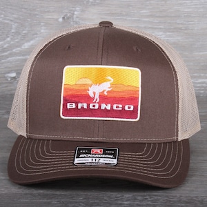 May include: A brown and tan trucker hat with a red, orange, and yellow embroidered patch that says "Bronco" and features a silhouette of a horse. The patch is surrounded by a white border. The hat has a black adjustable strap with a black label that says "Richardson 112 Original Trucker Adjustable".