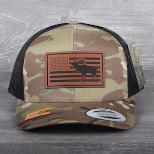 May include: A camouflage trucker hat with a black mesh back. The hat features a brown leather patch with an American flag and an elk silhouette. The text "The Authentic Snapback" is printed on the back of the hat.