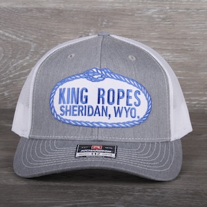 May include: Gray and white trucker hat with the text "KING ROPES SHERIDAN, WYO." in blue and white. The hat features a curved brim and a mesh back. The Richardson brand label is visible on the front.