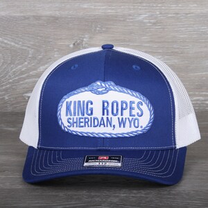 May include: A blue and white trucker hat with the text "KING ROPES SHERIDAN, WYO." embroidered on the front. The hat features a white mesh back and a curved brim. The hat has a Richardson 112 tag.